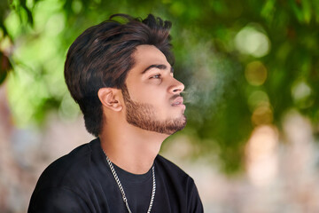 Attractive indian man smoker exhales cigarette smoke portrait in black t shirt and silver neck...