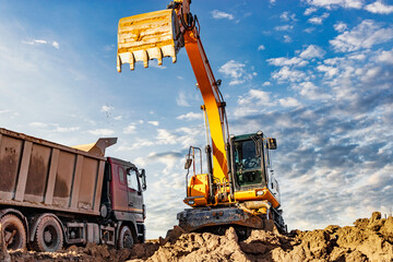 A wheeled excavator loads a dump truck with soil and sand. An excavator with a high-raised bucket against a cloudy sky View from the trench. Removal of soil from a construction site or quarry.