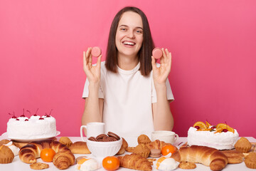Smiling pleased pretty brown haired woman in white t shirt sitting at table with desserts isolated over pink background holding cookies enjoying sweet dinner.