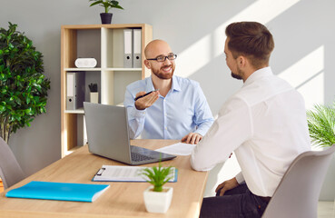 Business men at work in office. Two young serious employers sitting at the desk with laptop computer, talking and having discussion, considering new projects or startups, analyzing company.