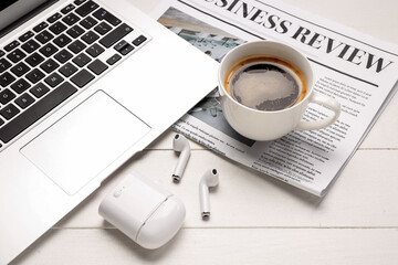 Cup of coffee with earphones, newspaper and laptop on white wooden background, closeup