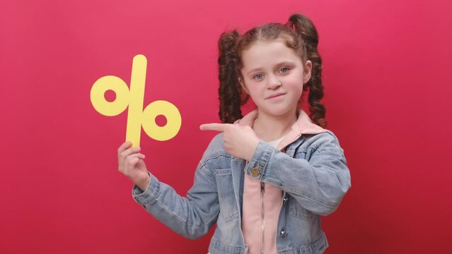 Portrait of pleasant cute little girl kid holding yellow percent mark, expressing positive emotions, presenting discounts, posing isolated over red color background wall in studio. Sale concept