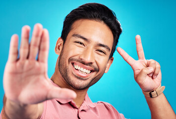 Happy asian man, portrait and peace sign for selfie, profile picture, or social media against a blue studio background. Male influencer or vlogger with smile showing peaceful emoji for photo or vlog