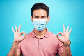 Man in face mask, portrait and corona with okay hand sign, safety from virus and bacteria isolated on blue background. Disease, covid and male person with emoji and health compliance in studio