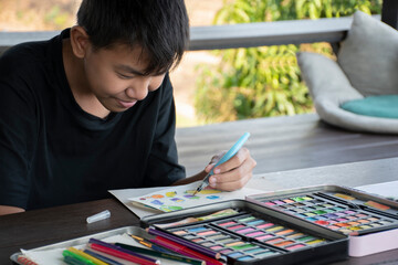 A boy in casual black clothes is practicing drawing and painting by using small watercolor rectangle bars in a paint box on a table on terrace of his summer vacation.