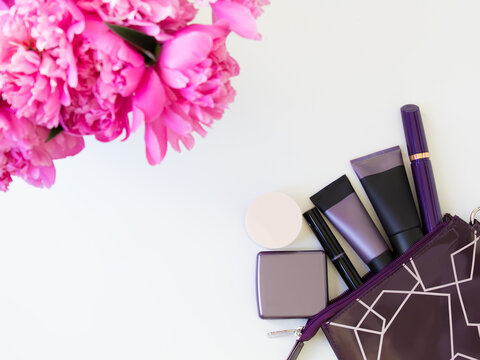 Glamorous purple black cosmetic set for women and men: foundation, lipstick, mascara, blush, powder, highlighter. Stylish cosmetics isolated on white background. Pink peonies in blur.