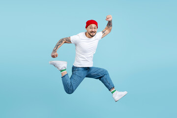 Fototapeta na wymiar Portrait of overjoyed asian man wearing red hat, white t shirt, stylish shoes jumping high, running, having fun isolated on blue background. Shopping, sales concept 