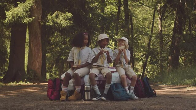 Three multiethnic elementary age kids in scout uniform eating homemade sandwiches and chatting in summer forest camp