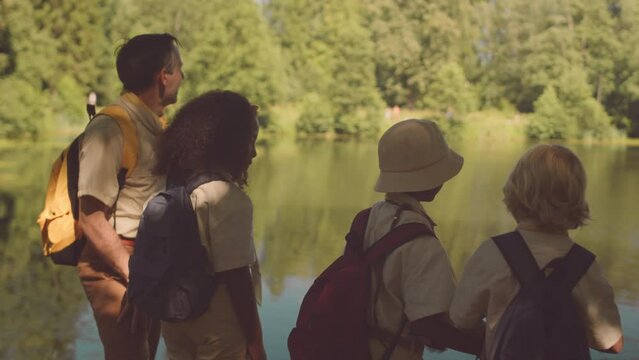 Back view of mid adult Caucasian male teacher and multiethnic kids in scout uniform looking at forest river through binoculars while scouting together in summertime