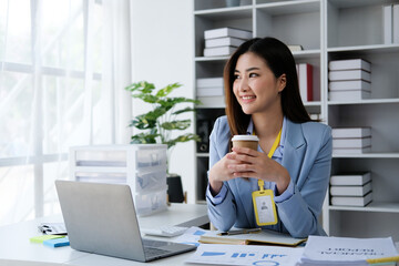 Asian businesswoman looking at camera drinking coffee working with laptop at office