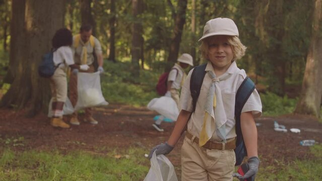 Medium portrait of little Caucasian boy scout with trash bag in hands posing for camera outdoors in forest, cleaning it from rubbish together with troop and their leader