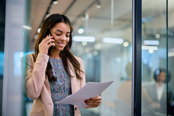 Fototapeta premium Happy black businesswoman talks on cell phone while reading documents in office.