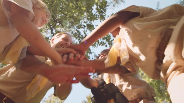 Low angle shot of cheerful diverse cub scouts and their leader putting their hands together standing in circle outdoors in summer forest