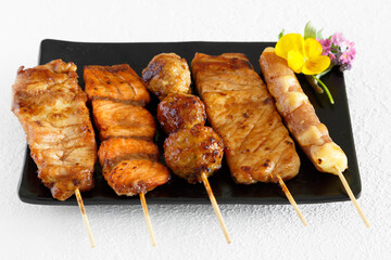 Assortiment de brochettes yakitori: brochette de poulet, brochette de boeuf fromage, brochette de saumon sur une assiette noir et fond blanc. Cuisine japonaise.