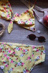 Floral yellow bikini, wicker bag, retro sunglasses, seashell, bag with peaches and cut watermelon on wooden background. Top view, copy space.