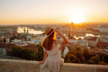 Smiling female tourist in a hat admires the landscape of the city at dawn. Euro-trip. Travel,...