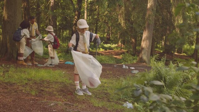 Full length shot of little Caucasian boy scout with troop of kids and their leader collecting rubbish in green forest together