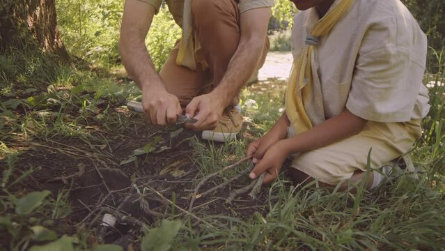 Adult Caucasian male scouter using knife and tree branch teaching young African American boy scout to make firewood while camping in forest