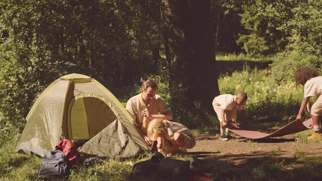 Diverse scout troop with leader setting up tent on forest glade while camping together at summertime