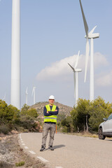 portrait of male person from afar with background of generators or windmills, wearing safety clothing plus helmet
