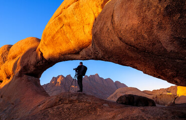 Nature photographer takes photo on his camera with tripod in a stone arch of the rock on Spitzkoppe, Namibia. Calm landscape at sunrise with silhouette of tourist in a mountain valley on Spitzkoppe. - 594567230