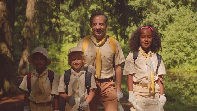 Medium portrait of scouter with diverse kids holding trash bags posing for camera in summer forest, helping to clean environment together