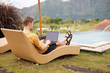 Young digital nomad working remotely on his laptop in Bali. Freelancer Man in workation video call. Travel and remote online work. Beautiful outdoor by the pool for remote workers someone abroad.