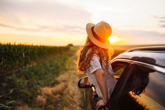 Young happy woman in hat enjoys car ride, leaning out of the window. In summer, a beautiful tourist woman travels by car and enjoys the golden sunset. Concept of an active lifestyle, travel, tourism.
