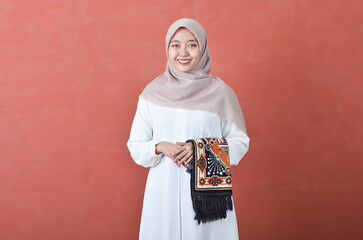 Beautiful Asian woman in white gamis and hijab smiling cheerfully buring prayer rugs isolated in brown background