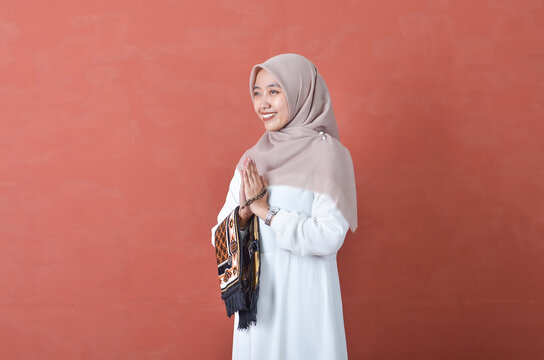 Beautiful Asian woman in white gamis and hijab smiling cheerfully namaste gesture and buring prayer rugs and prayer beads isolated in brown background