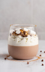 Chocolate mousse with whipped cream, marshmallows, chocolate drops and cinnamon in a glass. Portioned double-layer dessert on a marble background.