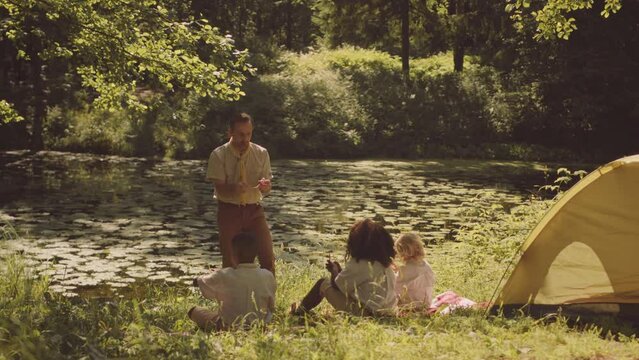 Mid adult Caucasian male scout leader teaching diverse group of kids camping skills sitting on green forest glade lit by sunlight