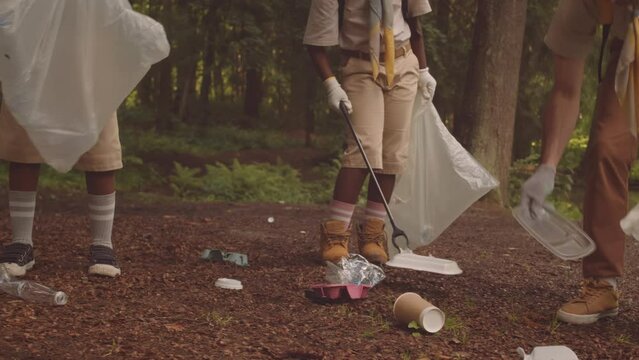 Diverse children from scout troop and their adult leader cleaning forest together picking up rubbish from ground and putting it into plastic trash bags
