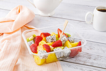 Fruit salad of pineapple, strawberries, kiwi and pitahaya in a plastic container on a wooden background.