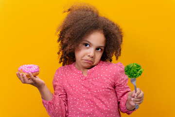 Studio shot of a confused Black girl holding fresh broccoli and donut on a yellow background.