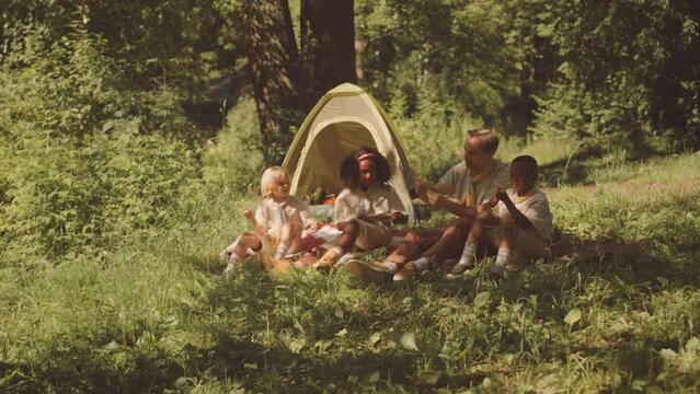 Multiethnic scout troop with leader sitting on forest glade by tent under sunlight learning to make ties