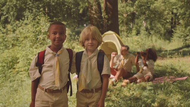 Medium slowmo portrait of two diverse elementary age boy scouts posing for camera standing against tent on sunlit forest glade, camping together with troop and their scout leader in summer