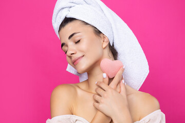 Beautiful cheerful attractive girl with a towel on her head, holds a sponge in the form of a pink heart.