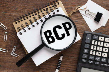 BCP Business Continuity Plan. wooden background with text on magnifier glass. two notepads and a calculator