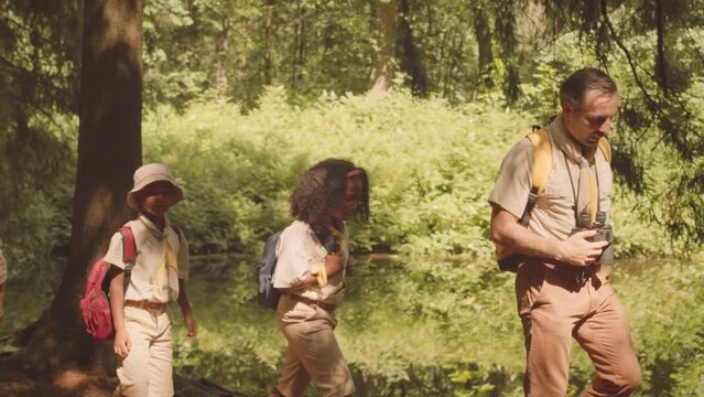 Diverse children in scout uniform following their leader while hiking in summer forest together