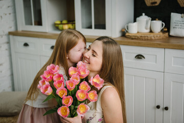 Happy birthday. Mom kiss daughter surprised. Kid congratulates mother and gives bouquet of tulip flowers in kitchen at home. Mother's Day concept. Greeting card. Women's Day. Back view. Family holiday