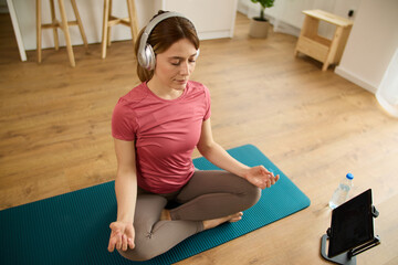 Young woman meditating at home while listening to tutorial lessons on digital tablet