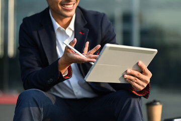 Pleased young businessman in formalwear sitting outside of office in urban city and using digital tablet
