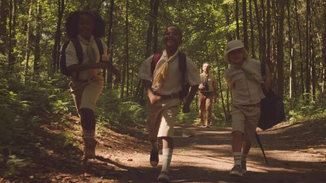 Slowmo of group of multiethnic cub scouts running along forest path under sunlight with their leader scout following them behind