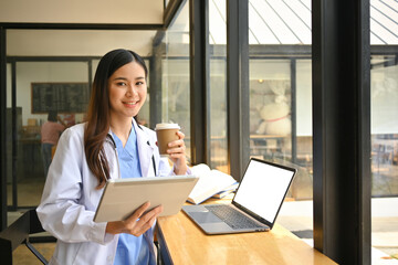 Fototapeta premium Positive female doctor in white coat having a break, drinking coffee and using digital tablet in hospital canteen