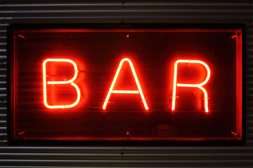 A bright red neon sign with the word bar in front of a striped metal wall at night. Luminous bar sign.