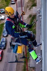 Fototapeta premium Professional industrial mountaineering worker washing exterior facade glazing building at urban background, hangs over house. Rope access laborer climbing on wall of skyscraper. Copy ad text space