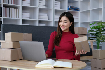 Smiling young Asian business owner woman prepare parcel box and standing check online orders of product for deliver to customer on laptop computer. Shopping Online concept.