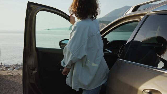 A young Caucasian woman gets out of her car parked by the sea and enjoys the beautiful sea view.