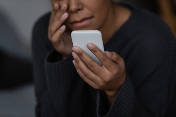 Cropped view of blurred multiracial woman with depression holding smartphone at home.